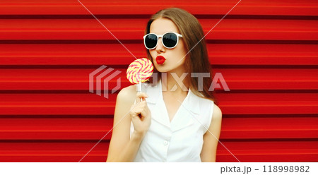 Portrait of stylish young woman blowing kiss with red lipstick wearing sunglasses on red background 118998982