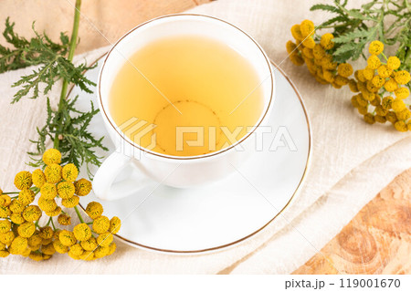 Healthy herbal tansy tea in white cup on saucer and tansy flowers on white napkin close up. Healthy herbal tansy tea in white cup on saucer and tansy flowers on white napkin close up. 119001670