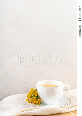 White cup on saucer with tansy decoction, tansy flowers on white napkin. Herbal medicine. Copy space White cup on saucer with tansy decoction, tansy flowers on white napkin. Herbal medicine. Copy space 119001671