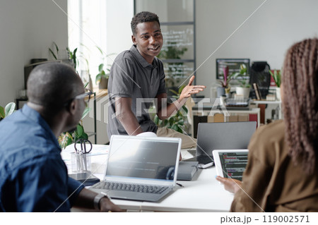Young African American an sitting on white table while telling joke during conversation with his coworkers in office Young African American an sitting on white table while telling joke during conversation with his coworkers in office 119002571