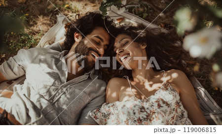 A couple lies on a blanket under a tree, laughing and pointing at cloud shapes as leaves rustle gently. A couple lies on a blanket under a tree, laughing and pointing at cloud shapes as leaves rustle gently. 119006815