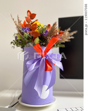 Colorful bouquet of flowers in a lavender box standing on a desk, tied with a vibrant red and purple ribbon. Bright, mixed arrangement adds a touch of nature and warmth to modern workspace 119007086