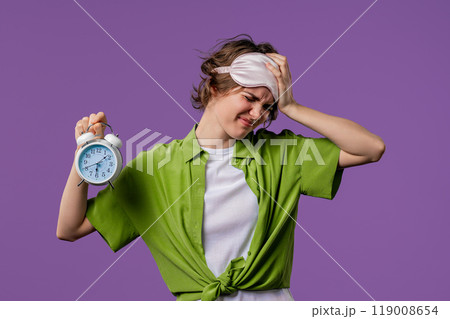 Woken up by alarm clock sleepy young woman holding it in hand. Violet background. Early 6 o'clock in morning. Lazy lady didn't get enough sleep, concept of passing time. 119008654