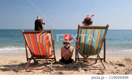 Young family of three in festive hats relaxes on the beach, enjoying colorful fruit cocktails and soaking up the sun, celebrating Christmas and New Year Young family of three in festive hats relaxes on the beach, enjoying colorful fruit cocktails and soaking up the sun, celebrating Christmas and New Year 119008996