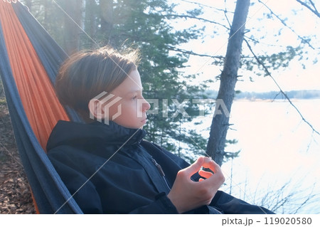 Teenager boy relaxing lying in hammock eating sausage in forest on nature. 119020580