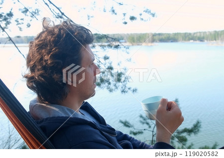 Curly man relaxing lying in hammock drinking tea in forest admiring blue lake. 119020582