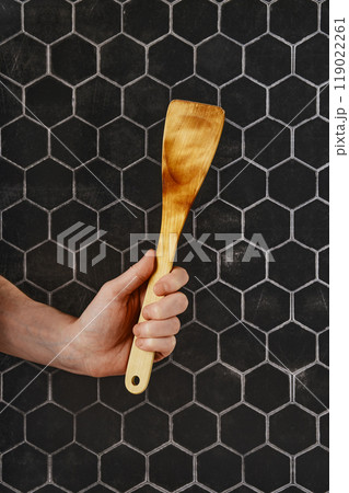 A hand holding a wooden spatula against a black hexagonal tile background in a kitchen setting 119022261