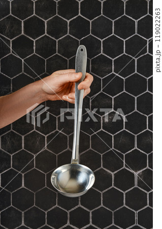 A hand holding a long-handled ladle against a hexagonal tile background in a kitchen setting 119022263