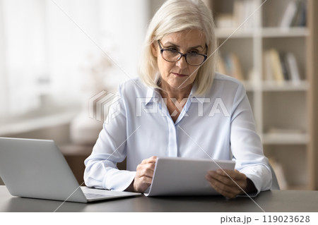 Older woman reading document seated at desk with laptop Older woman reading document seated at desk with laptop 119023628