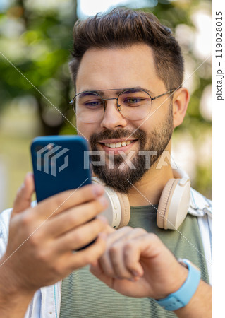 Smiling young man using smartphone typing texting scrolling social media app on city town street 119028015