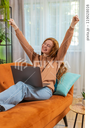 Excited woman sitting on sofa using laptop receives good news victory celebrate lottery win at home 119028016