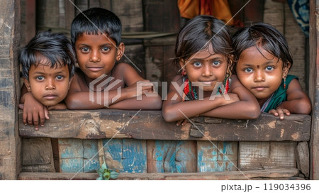 Four curious children with big eyes peer out from rustic window in their Indian village home. 119034396