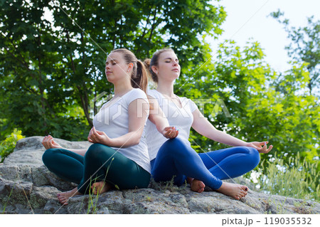 Two healthy calm young women meditating in nature with eyes closed, doing breathing exercises, relaxing body and mind. 119035532