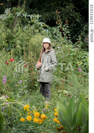 Girl wearing a jacket and a white hat walks through the garden with flowers holding a pumpkin 119035818