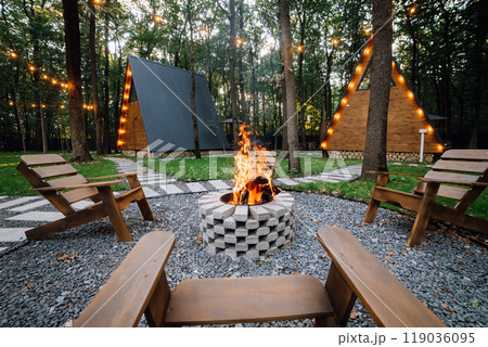 Campfire area with wooden chairs and cabins in background, creating a cozy outdoor gathering space Campfire area with wooden chairs and cabins in background, creating a cozy outdoor gathering space 119036095