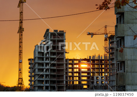 Golden sunset behind a bustling construction site with cranes and buildings silhouetted against the vibrant sky 119037450