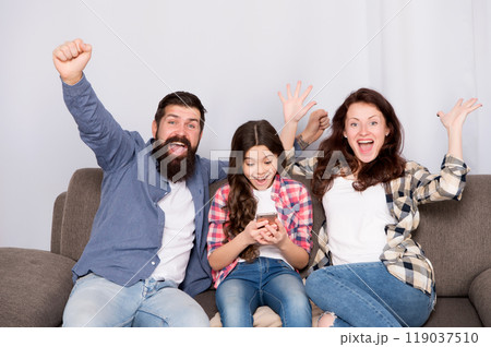 Child little girl use smartphone with parents. Friendly family having fun together. Mom dad and busy daughter relaxing on couch. Family leisure. Parental advisory. Family spend weekend together 119037510