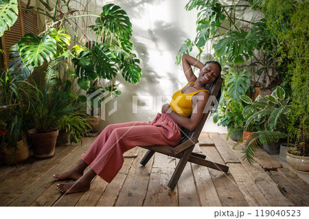 Happy african american woman smiling enjoy break relaxed sits on chair in urban jungle sunny room. Happy african american woman smiling enjoy break relaxed sits on chair in urban jungle sunny room. 119040523