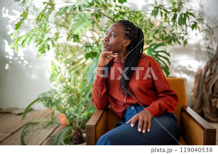 Thoughtful african american woman sitting on armchair on houseplants background pondering look aside 119040538