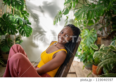 Pleased relaxed black woman looking at camera rest on cozy chair surround by tropical indoor plants. Pleased relaxed black woman looking at camera rest on cozy chair surround by tropical indoor plants. 119040597