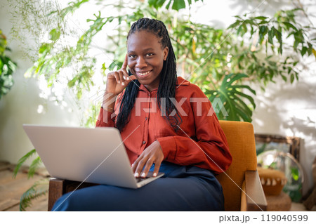 Happy pleased black woman with laptop on laps sitting on chair in sunny room smiling look at camera. 119040599