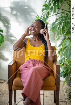 Happy black woman relaxing in armchair in cozy apartment with houseplant, listen music in headphones Happy black woman relaxing in armchair in cozy apartment with houseplant, listen music in headphones 119040636