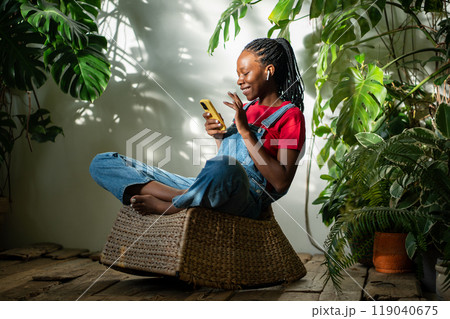 Cheerful black woman relaxing at indoor garden. African American with smartphone among sunny garden Cheerful black woman relaxing at indoor garden. African American with smartphone among sunny garden 119040675