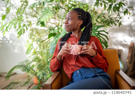 Dreaming african american woman holding cup resting on armchair surround houseplants looking aside. 119040745