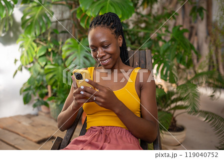 Joyful black woman smiling using smartphone relaxing chatting in social media sit among houseplants. Joyful black woman smiling using smartphone relaxing chatting in social media sit among houseplants. 119040752