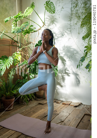 Black woman meditating in yoga tree pose stand on one leg with closed eyes at home garden. Wellness. 119040781