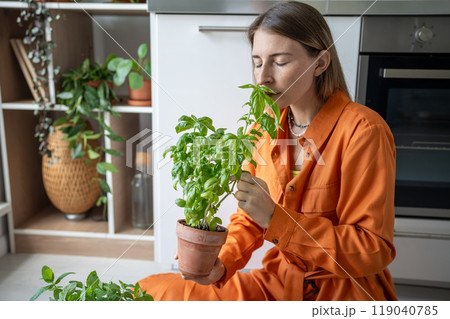 Woman enjoying smell of basil. Girl sits on floor, inhales aroma of home-grown flavour basil in pot 119040785