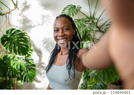 Happy african american woman smiling making selfie on smartphone surround by tropical houseplants. 119040801