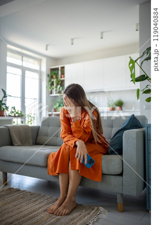 Unhappy sad woman in difficult situation sitting at home on couch with smartphone received bad news Unhappy sad woman in difficult situation sitting at home on couch with smartphone received bad news 119040884