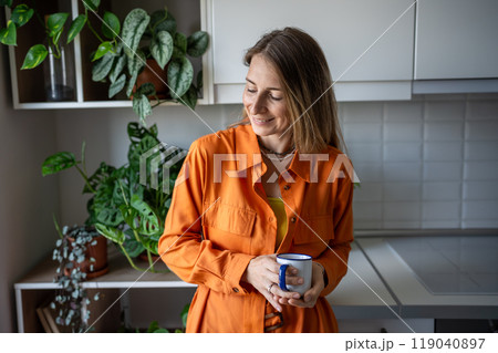 Joyful woman smiling standing in greenery kitchen with cup of coffee planning good day in morning Joyful woman smiling standing in greenery kitchen with cup of coffee planning good day in morning 119040897