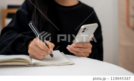 A close-up of a woman writing in a book and using her smartphone while sitting at a table indoors. 119045387