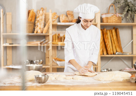 Young woman works as baker, mold baguette from dough, creates long loafs from dough. Young woman works as baker, mold baguette from dough, creates long loafs from dough. 119045451