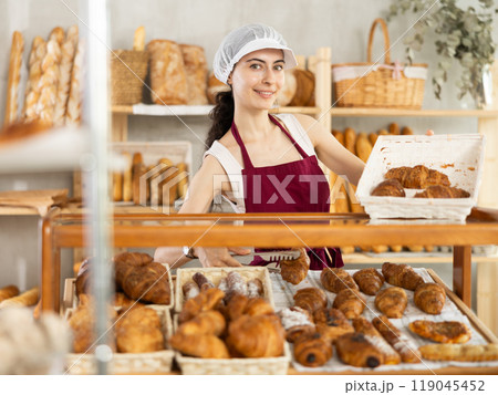 Young woman employee puts croissants in window, arranges display of goods at bakery. Young woman employee puts croissants in window, arranges display of goods at bakery. 119045452