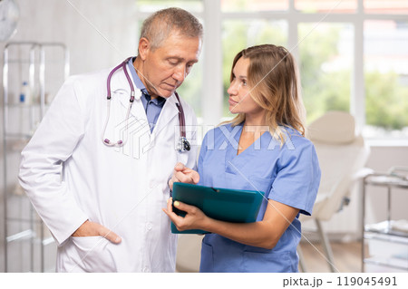 Middle-aged nurse and mature doctor discussing diagnosis standing in cabinet Middle-aged nurse and mature doctor discussing diagnosis standing in cabinet 119045491