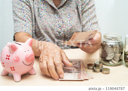 Asian elderly woman putting coin into pink piggy bank for saving money and insurance, poverty, financial problem in retirement. 119048787