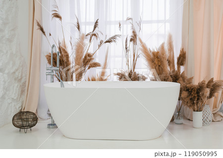 White bathtub with a faucet and a window behind it, decorated dried grasses 119050995