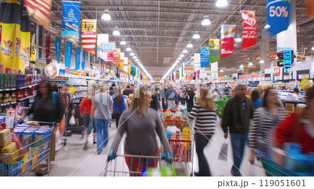 Blurred motion of people in supermarket. Wide-angle shot of a busy store with colorful sale banners hanging from the ceiling. Shopping carts filled with products are in the foreground. 119051601