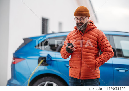 Man charging electric car during cold snowy day, using electric vehicle charging app, checking battery life, energy consumption on smart phone. Charging and driving electric vehicles during winter Man charging electric car during cold snowy day, using electric vehicle charging app, checking battery life, energy consumption on smart phone. Charging and driving electric vehicles during winter 119053362