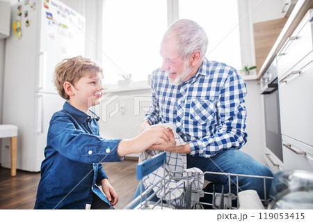 Boy helping grandfather clean the kitchen, unloading clean dishes out of the dishwasher. Grandfather taking care of grandson while parents are at work. Boy helping grandfather clean the kitchen, unloading clean dishes out of the dishwasher. Grandfather taking care of grandson while parents are at work. 119053415