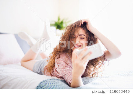 Portrait of beautiful young woman with curly hair lying on bed and taking selfie. Portrait of beautiful young woman with curly hair lying on bed and taking selfie. 119053449