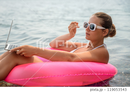 A woman is sitting in a pink inflatable raft on a lake, holding a laptop. Concept of relaxation and leisure, as the woman enjoys her time in the water while working on her laptop. A woman is sitting in a pink inflatable raft on a lake, holding a laptop. Concept of relaxation and leisure, as the woman enjoys her time in the water while working on her laptop. 119053543