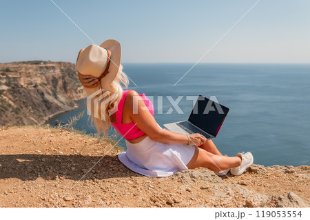 A woman is sitting on the beach with a laptop open in front of her. She is wearing a pink top and a straw hat. Concept of relaxation and leisure. 119053554