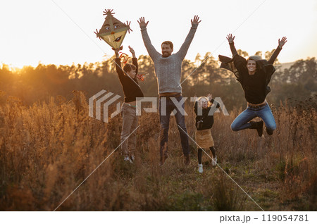 Portrait of young family flying a kite in the autumn nature, jumping in the middle meadow during sunset. Indian summer. Portrait of young family flying a kite in the autumn nature, jumping in the middle meadow during sunset. Indian summer. 119054781