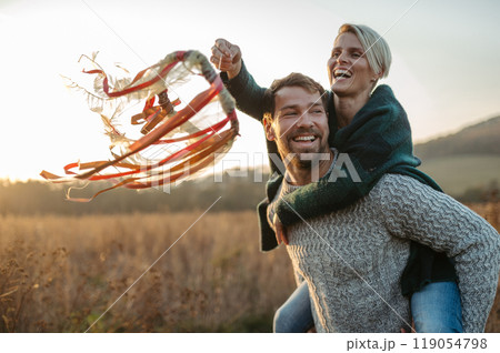 Couple in love on a walk in autumn forest. Husband piggycarrying wife on back, holding wreath with ribbons. Couple in love on a walk in autumn forest. Husband piggycarrying wife on back, holding wreath with ribbons. 119054798