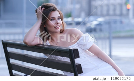 Carefree beautiful woman in white summer dress sitting on bench on city street on summer evening 119055176