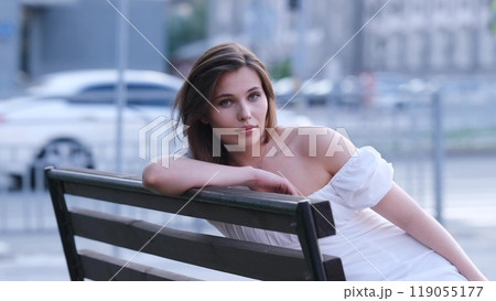 Carefree beautiful woman in white summer dress sitting on bench on city street on summer evening 119055177